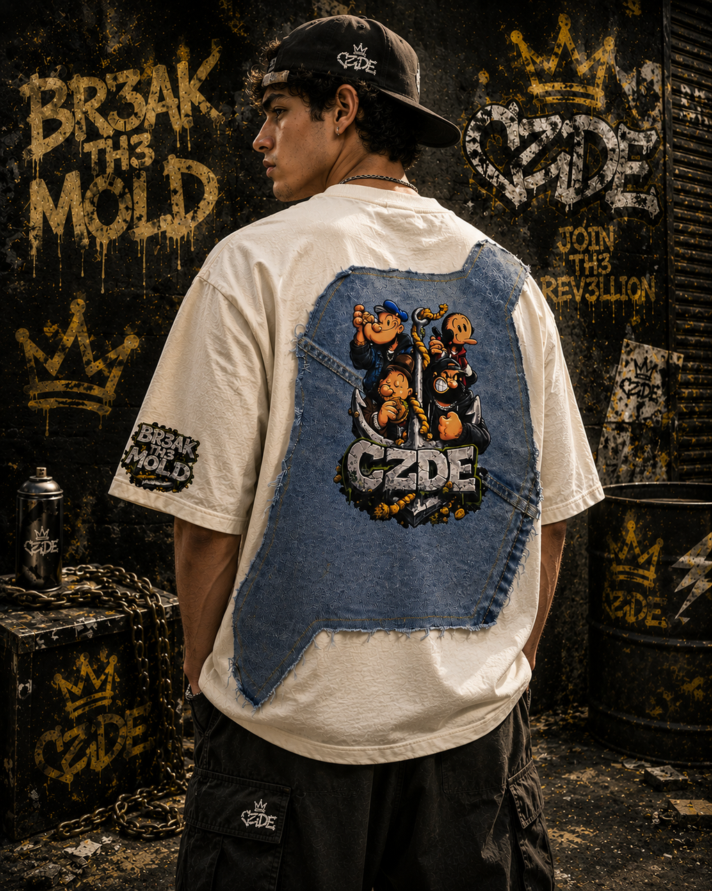 Male model wearing CZDE x Popeye Denim Patch Tee – back view showing full Popeye crew denim panel, BR3AK TH3 MOLD sleeve patch, JOIN TH3 R3V3LLION graffiti wall editorial